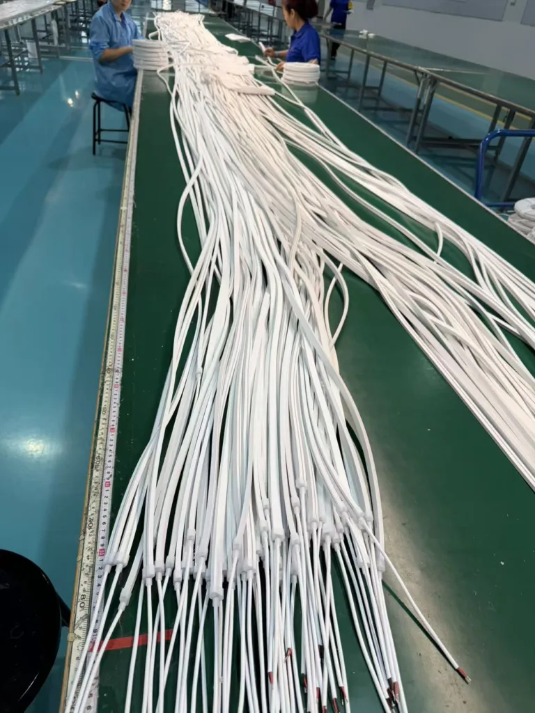 Rows of white silicone neon flex lights organized on a green workbench inside a factory, with workers assembling components.