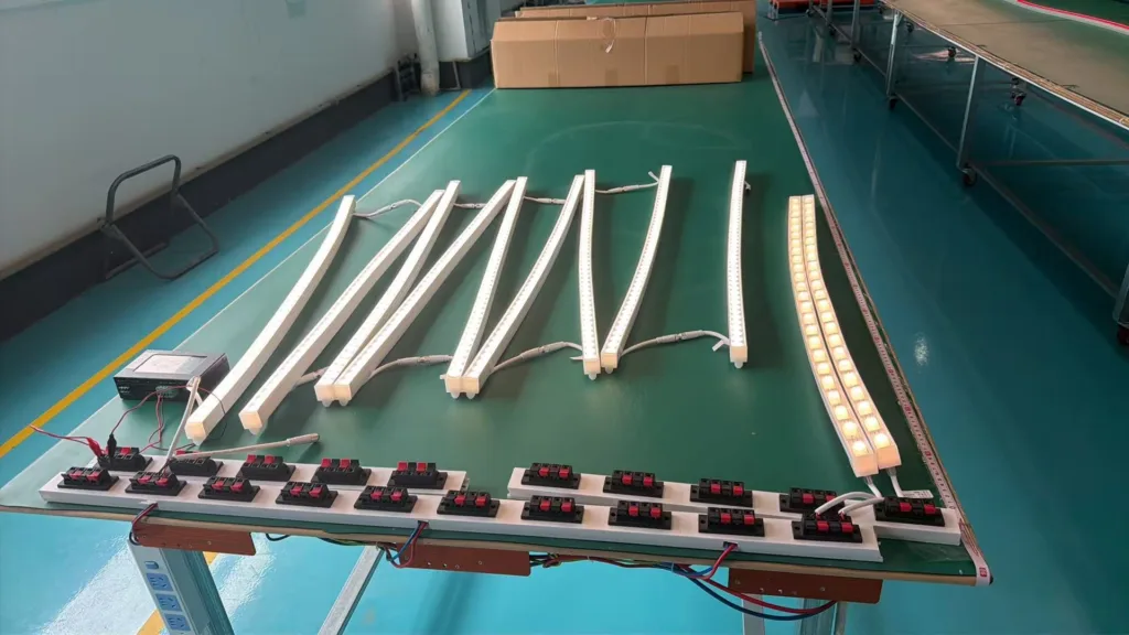 Curved and straight silicone LED lights on a testing bench with wiring and connectors in a factory setting.