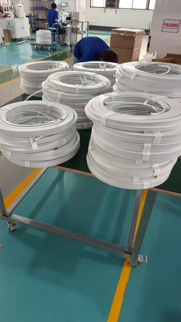 Rolls of silicone neon flex lighting material stacked on a table in a manufacturing facility with workers and machinery in the background.