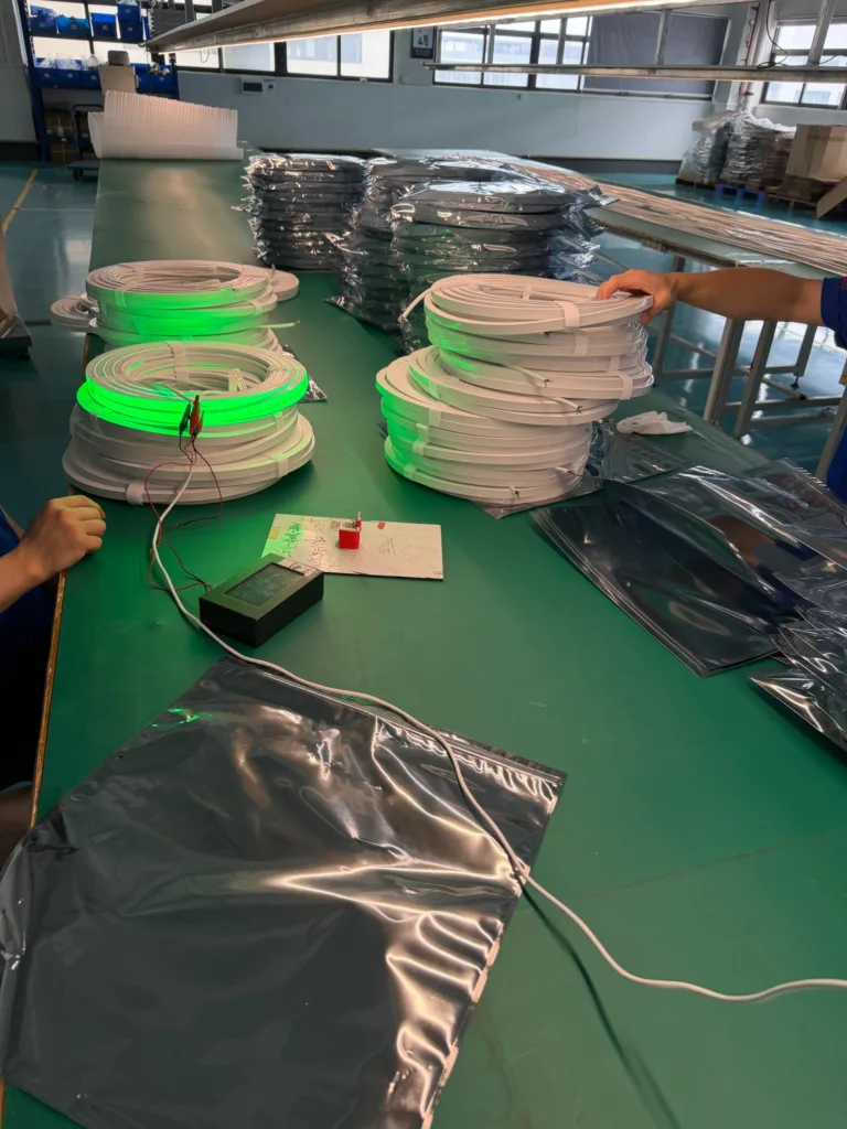 Factory workbench with silicone neon flex lights being tested, displaying green illumination on coiled strips with workers handling materials.