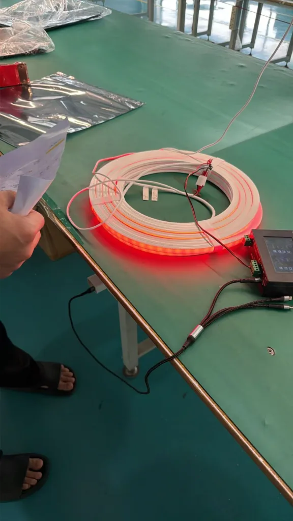Coiled silicone neon flex emitting red light connected to testing equipment on a workbench with a technician holding a document nearby.