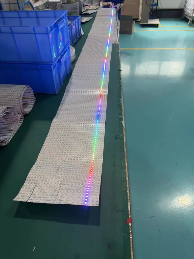 LED light strips being tested in a manufacturing facility, on a green workbench with multi-color illumination and nearby plastic bins.