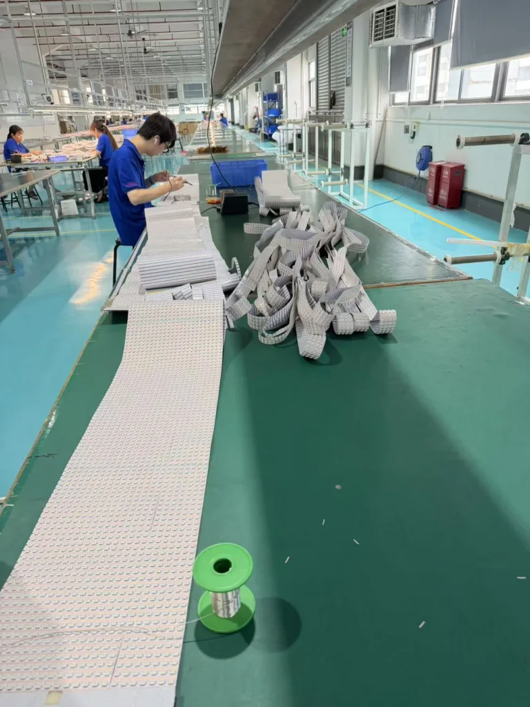 Factory workers assembling LED lighting components in a clean, organized workspace with green workstations and extrusion materials.