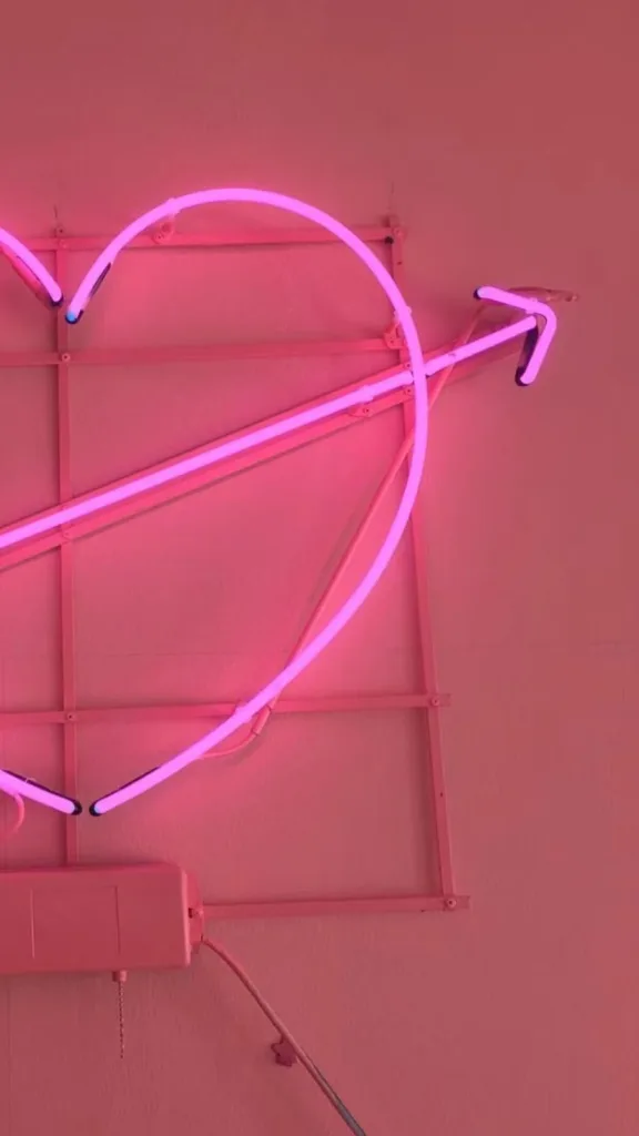 Bright pink neon lighting shaped as a heart with an arrow passing through, mounted on a pink wall for decorative display.