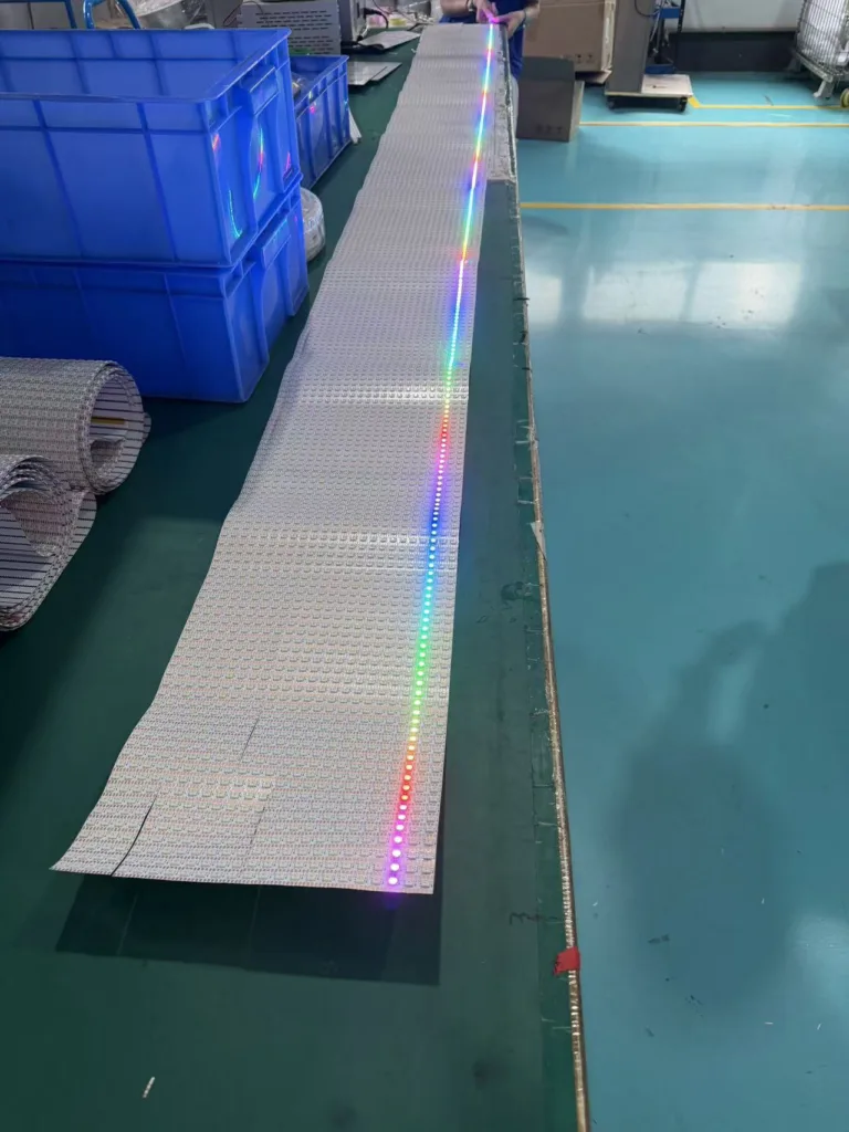 Long white LED panel strip emitting RGB lights on a production line with blue bins and industrial workspace background.
