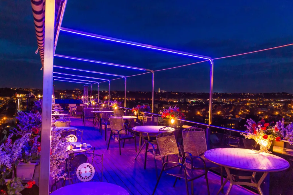 Outdoor rooftop seating area illuminated with purple LED lights, featuring tables and chairs, floral decor, and a cityscape view at night.