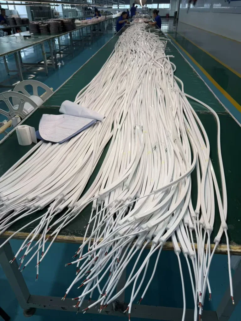 Rows of silicone neon flex cables on a production line table inside a manufacturing facility, with workers assembling parts in the background.