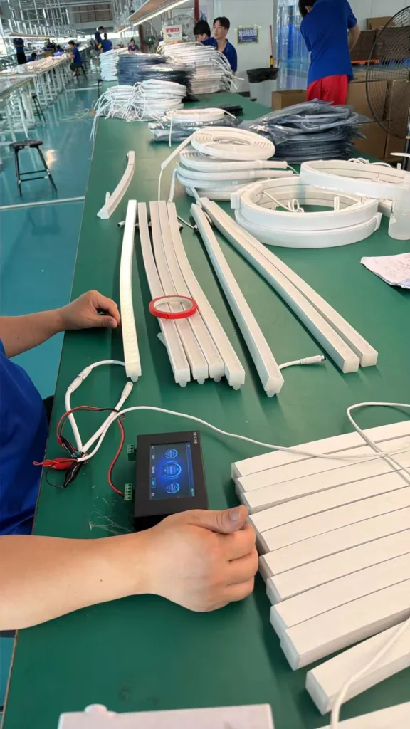 Worker assembling silicone neon lights on a production line with various lighting components and a control device on a green worktable.