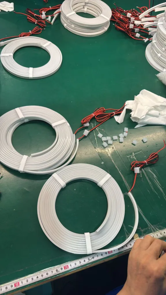 Coiled silicone neon flex lights with red wiring laid on a green work surface, accompanied by connectors and a hand measuring with a ruler.