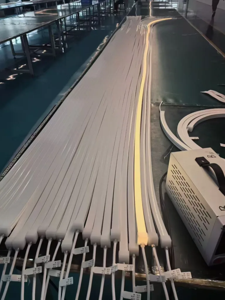 Silicone neon flex strips lined up on a production table with one illuminated strip and connected wires in a factory setting.